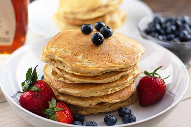 Horizontal image of a stack of flapjacks on a white dish with fresh strawberries and blueberries, with a bowl of blueberries and syrup in the background.