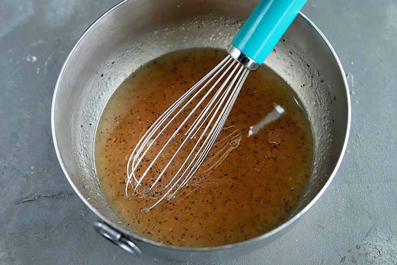 Horizontal image of a metal bowl with a light brown vinaigrette stirred by a whisk.