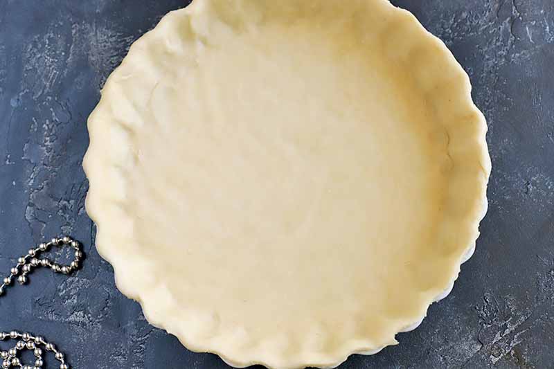 Overhead horizontal image of a prepared tart crust that's ready to be baked, with a beaded chain at the bottom left of the frame, on a dark gray surface.