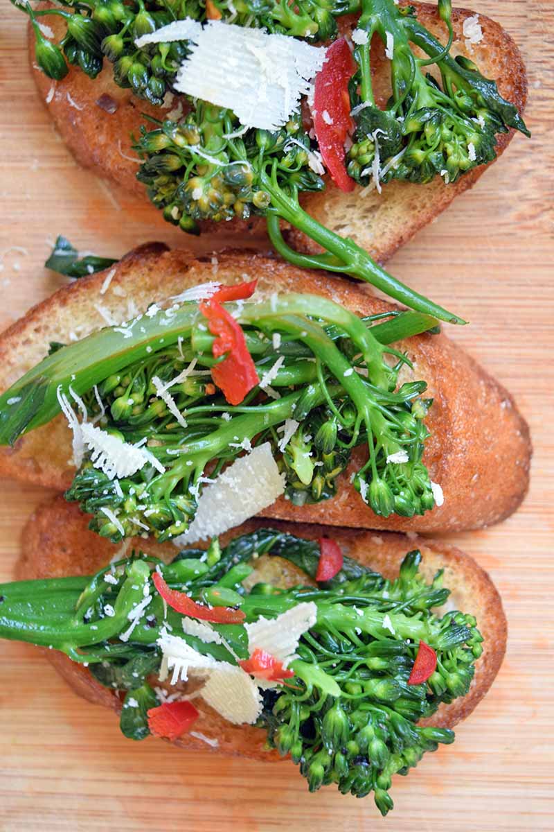 Vertical overhead image of three broccoli rabe tartines with thinly sliced red chili pepper and shaved aged hard cheese, on a light brown wood surface.