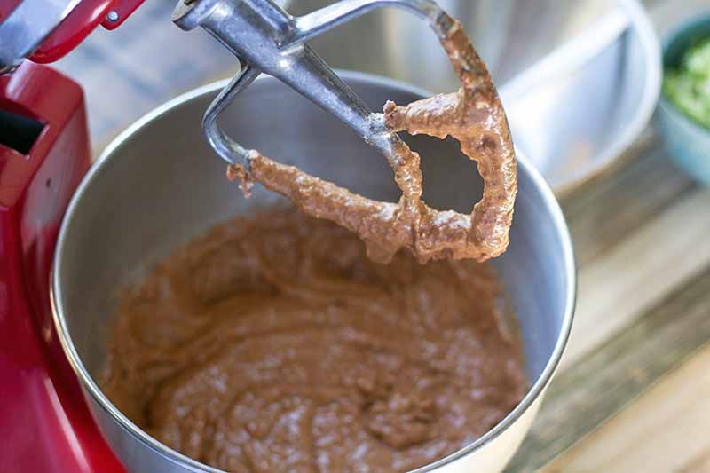 Horizontal image of chocolate batter on a paddle attachment beater and in a stainless steel bowl on a red stand mixer, with a white cloth and a small bowl of shredded zucchini in soft focus in the background, on a wood surface.