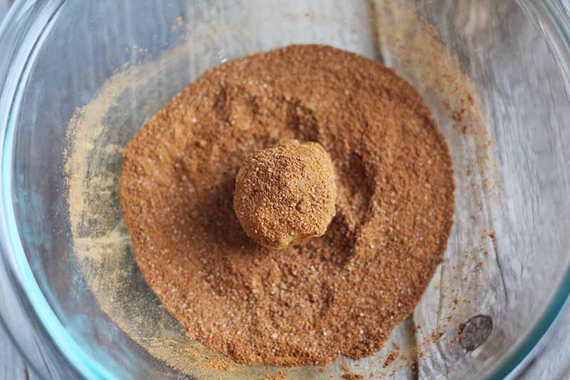 Horizontal overhead image of a large glass mixing bowl with a mixture of cinnamon and granulated sugar at the bottom, with a small ball of dough coated in the mix.