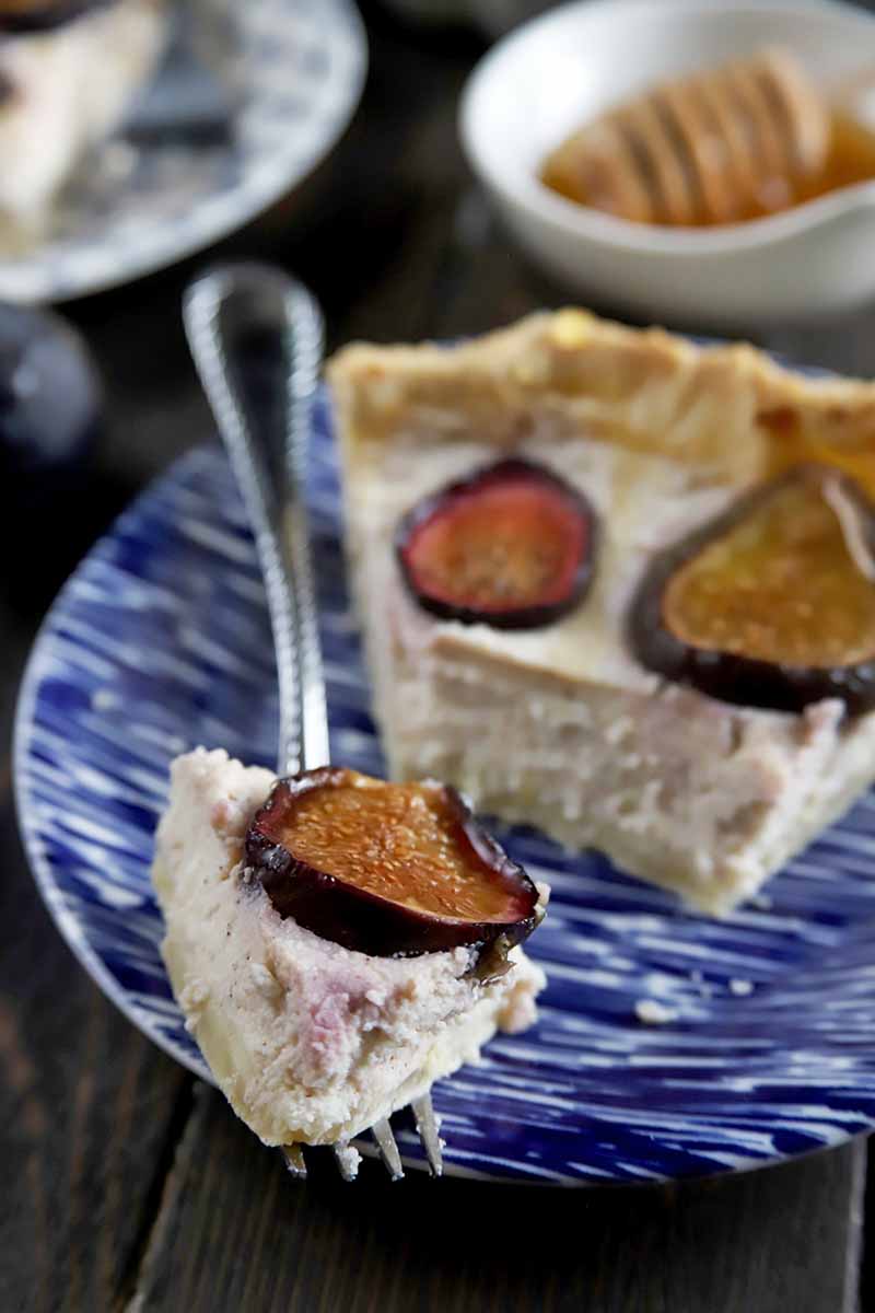 A bite of fig and ricotta tart on a fork in the foreground, on a blue and white plate with the remainder of the slice, with another plate and a small white dish of honey with a wooden dipper in soft focus in the background, on a dark brown wood surface.