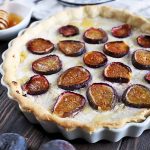 Horizontal image of a fig and ricotta tart in a white ceramic pan, on a dark brown wood table with a metal and wood pie server, a white dish of honey with a wooden dipper, and a few pieces of fresh fruit.