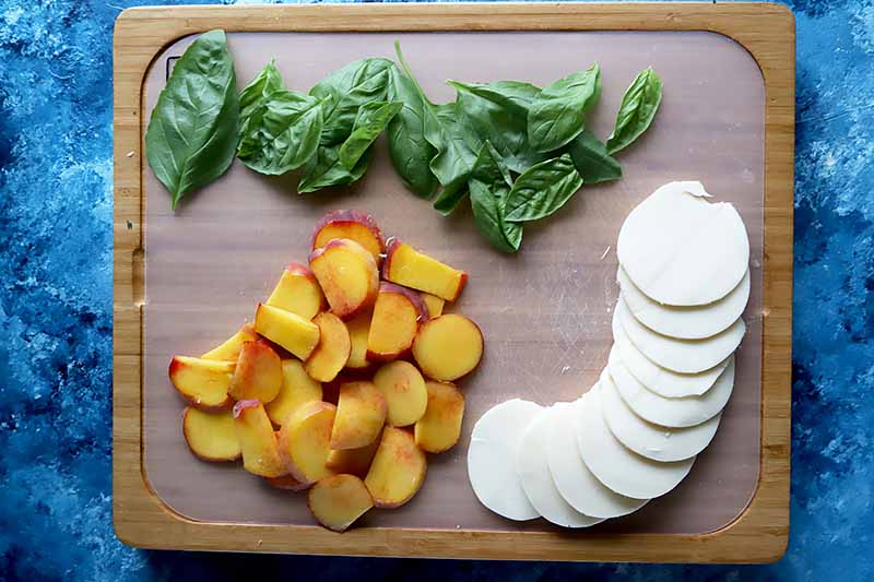 Horizontal image of a wooden cutting board with basil, peaches, and cheese.