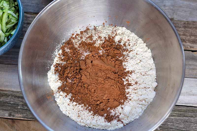 Horizontal overhead image of a stainless steel bowl of flour and cocoa powder, with a smaller blue ceramic bowl of shredded zucchini to the left, on an unfinished wood surface.