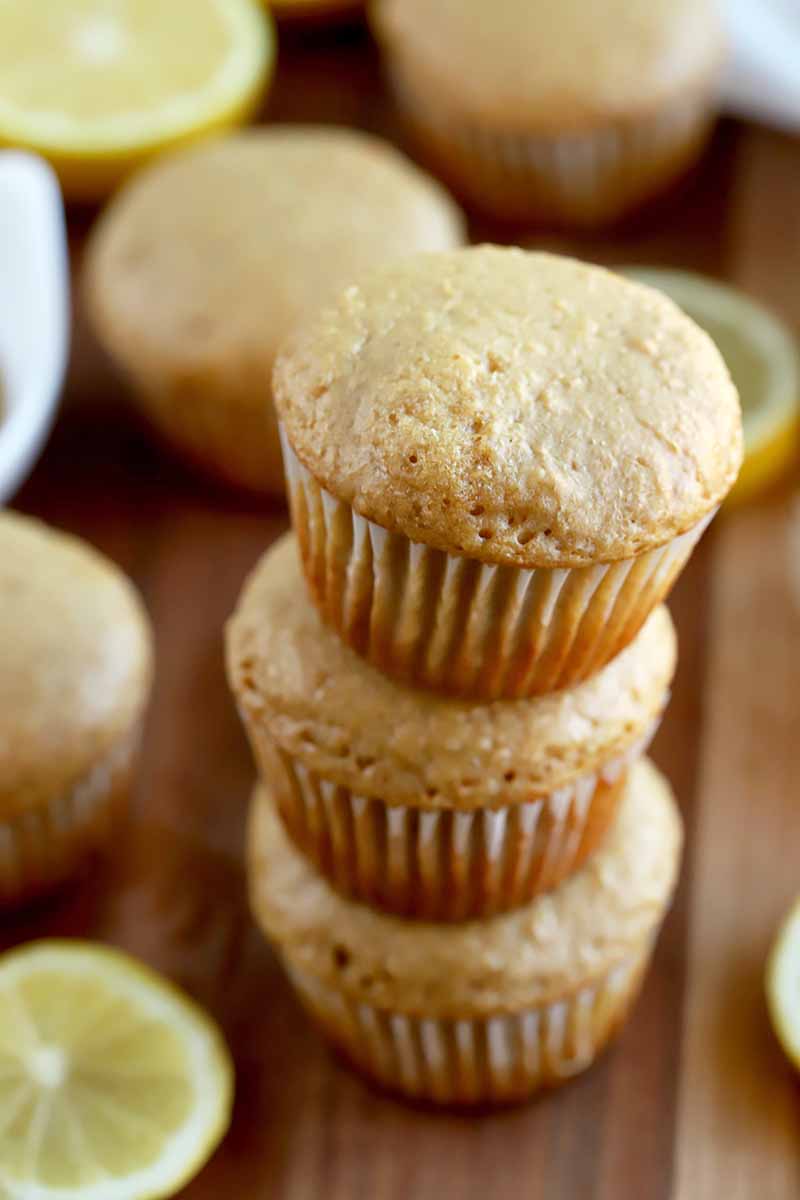 Vertical image of a stack of three muffins in paper wrappers, with more of the baked goods and scattered lemon slices on a brown wood surface.
