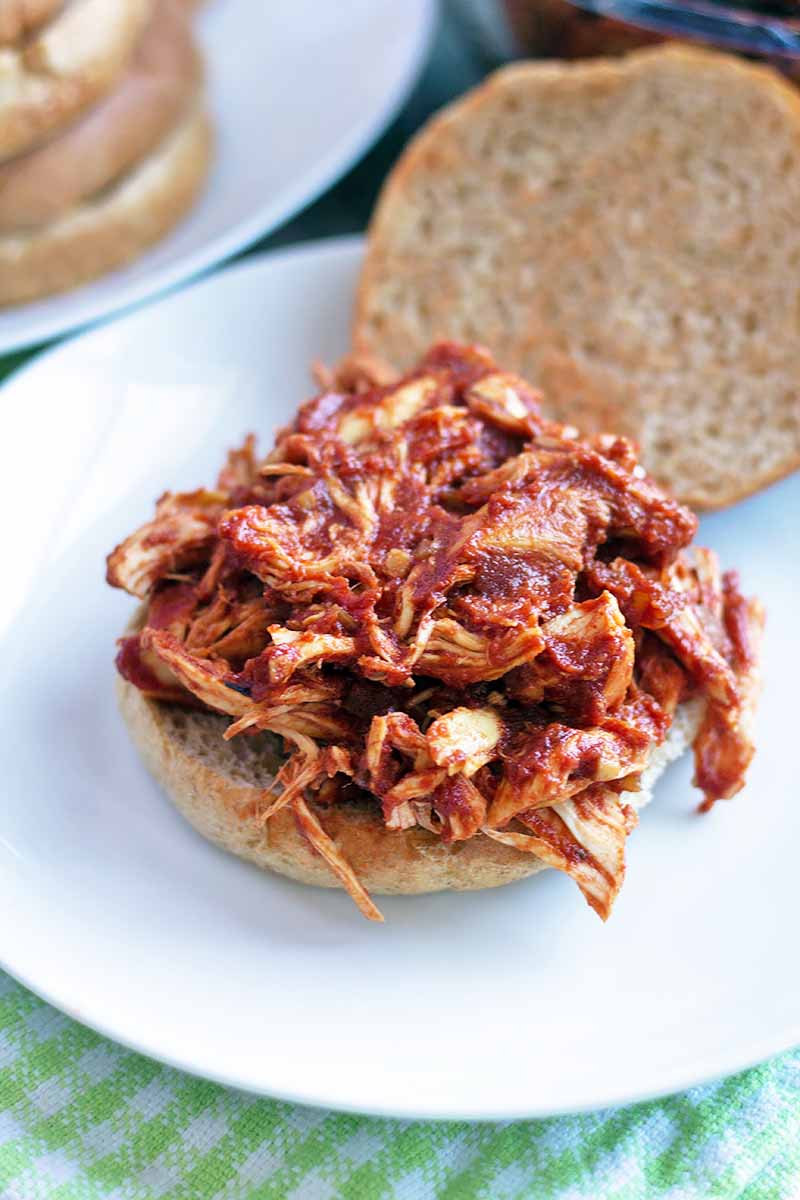 Vertical image of an open-faced shredded chicken sloppy joe sandwich on a white ceramic plate, with more buns on another plate in the background, on a pastel green and white checkered cloth.