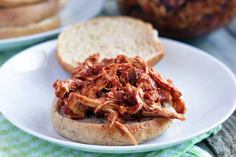 Horizontal image of shredded chicken breast in red sauce on a hamburger bun, on a white plate on top of a light green and white cloth on an unfinished wood table.