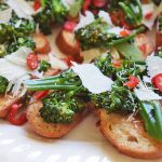 Horizontal image of a white platter of broccoli rabe tartines with sliced red chili and shaved hard cheese.