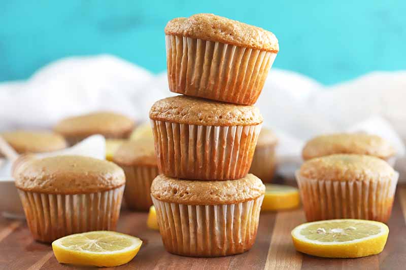 Horizontal image of a stack of three muffins surrounded by several more, with scattered lemon slices on a brown surface, with a white cloth in the background, against a sky blue backdrop.