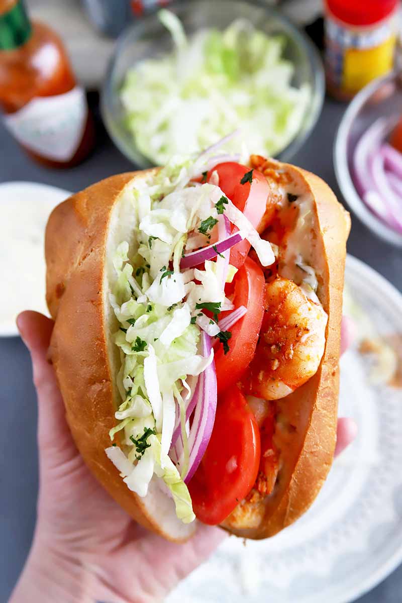 Vertical image of a hand holding a huge shrimp sandwich with fresh lettuce, red onions, and tomatoes.