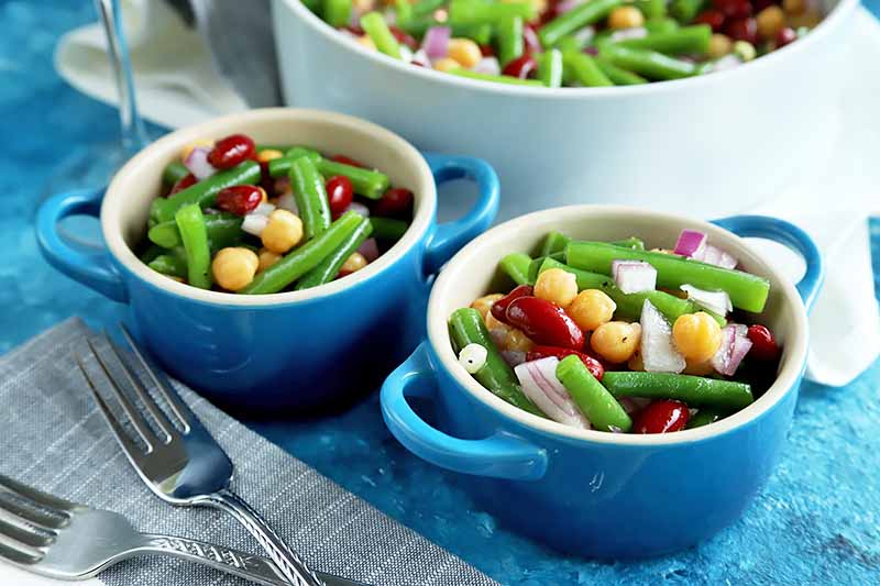 Horizontal image of two blue bowls filled with a colorful legume salad next to silverware.