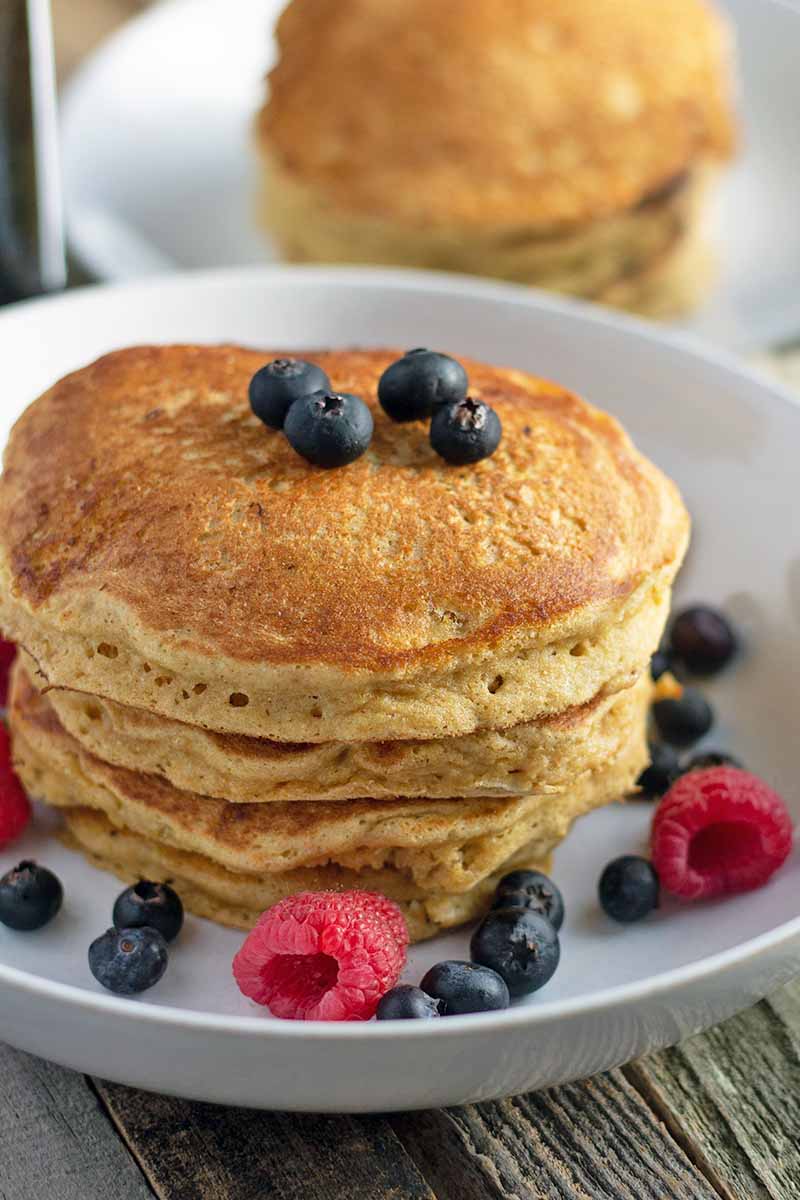 Vertical image of two white plates of pancakes, one in soft focus in the background, and one in sharp focus in the foreground, with scattered blueberries and raspberries, on a wood surface that has not been stained or varnished.