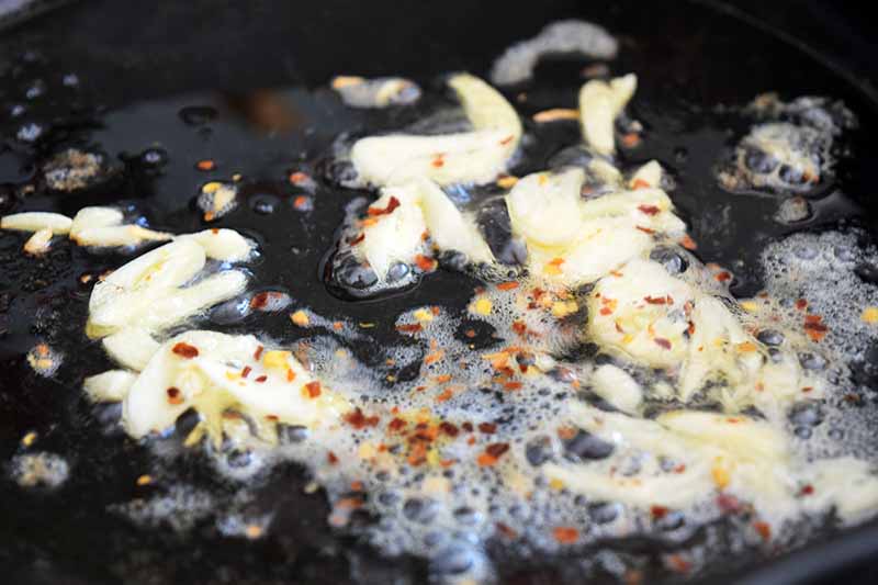 Closeup image of thinly sliced garlic and red chili pepper flakes being sauteed in a cast iron frying pan with foamy butter and olive oil.