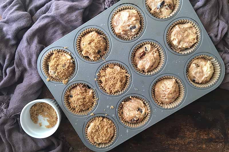 Horizontal image of batter in a muffin pan topped by brown sugar.