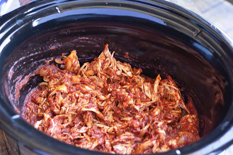 Oblique overhead image of shredded chicken in a red tomato-based sauce in a slow cooker.