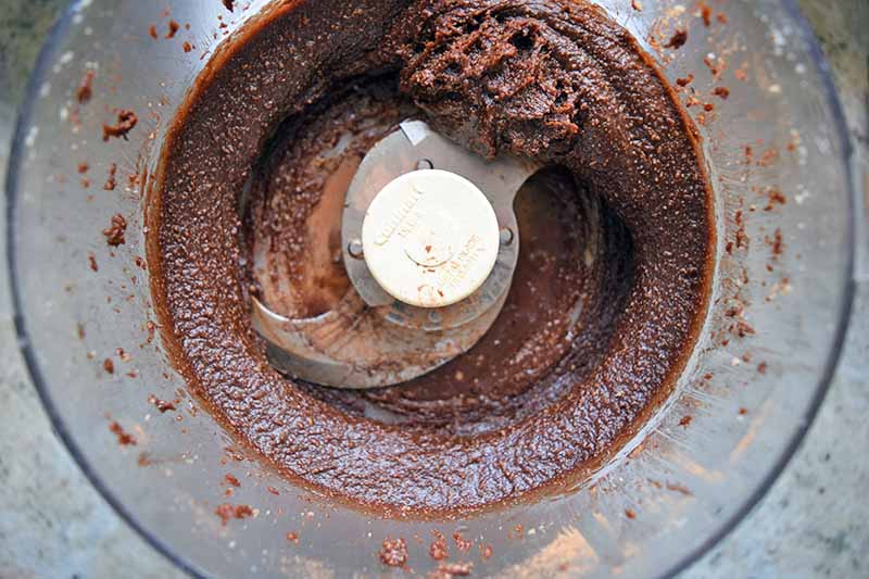 Horizontal overhead image of a chocolate mixture in the clear plastic canister of a food processor, with a metal S-blade attachment.
