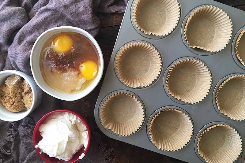 Horizontal image of a muffin pan with paper lines, a bowl of eggs, and a cup of yogurt.