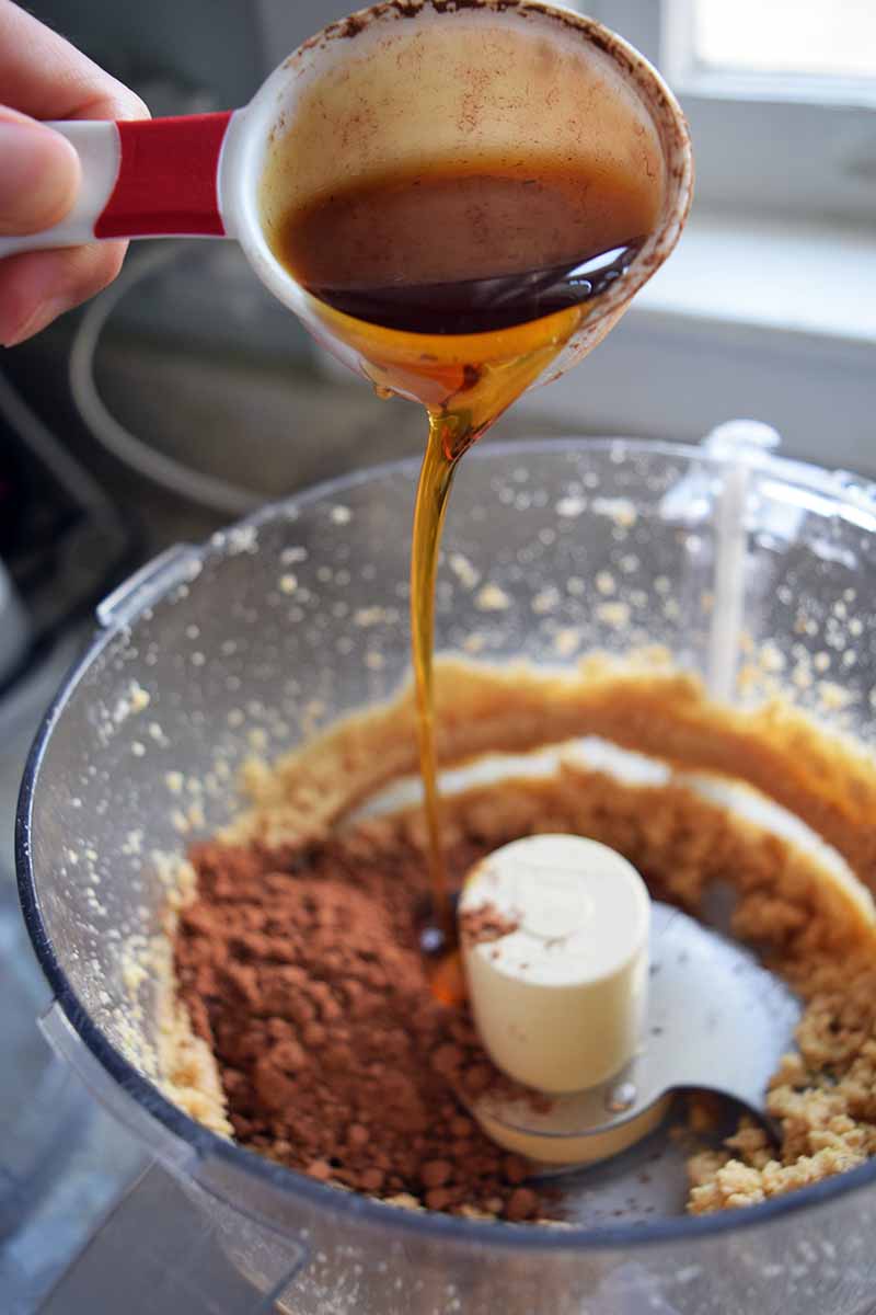 Vertical image of a hand pouring a measuring cup of maple syrup into a mixture of nut puree, cocoa powder, and other ingredients in a food processor below, on a gray kitchen countertop.