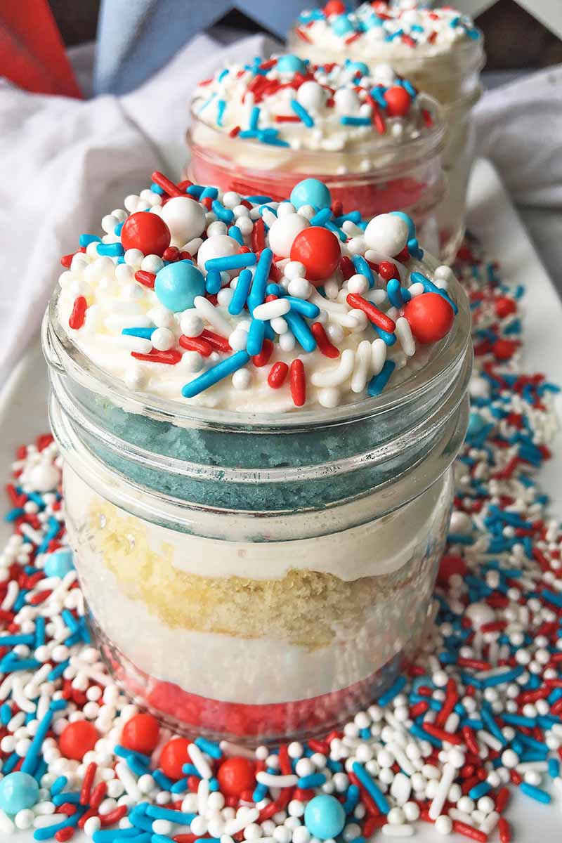Vertical image of glass containers with red, white, and blue cake circles and buttercream topped with assorted sprinkles.