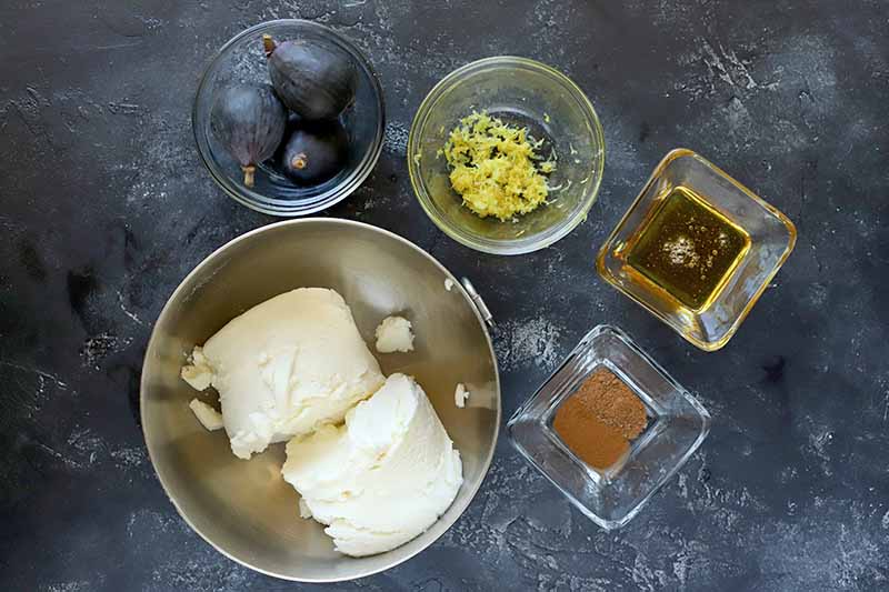Overhead image of a stainless steel bowl of ricotta with two small glass bowls of fresh figs and lemon zest, and two small square glass bowls of honey and warming spices, on a mottled dark gray background.