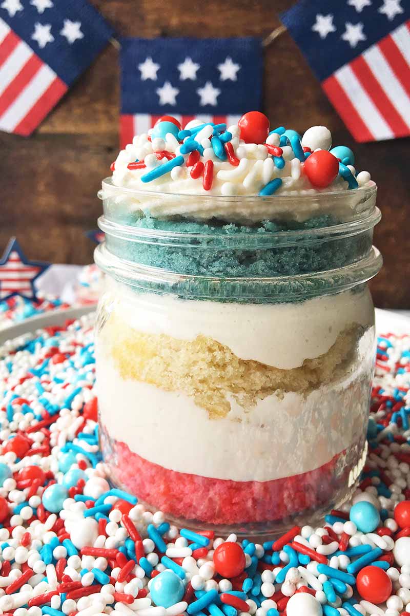 Vertical image of a glass jar with layers of red, white, and blue cake and white frosting surrounded and topped with sprinkles in front of flags.