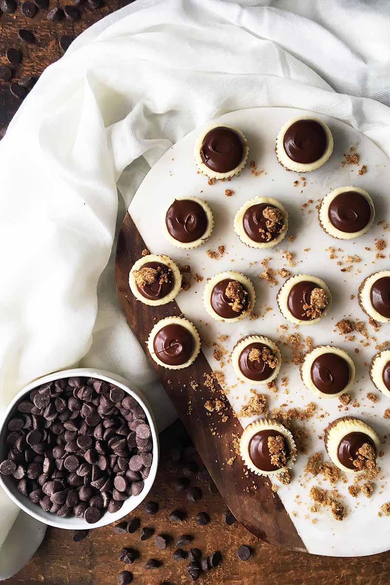 Vertical top-down image of a marble and wooden platter with chocolate-topped mini desserts next to a bowl of chocolate chips and a white towel.