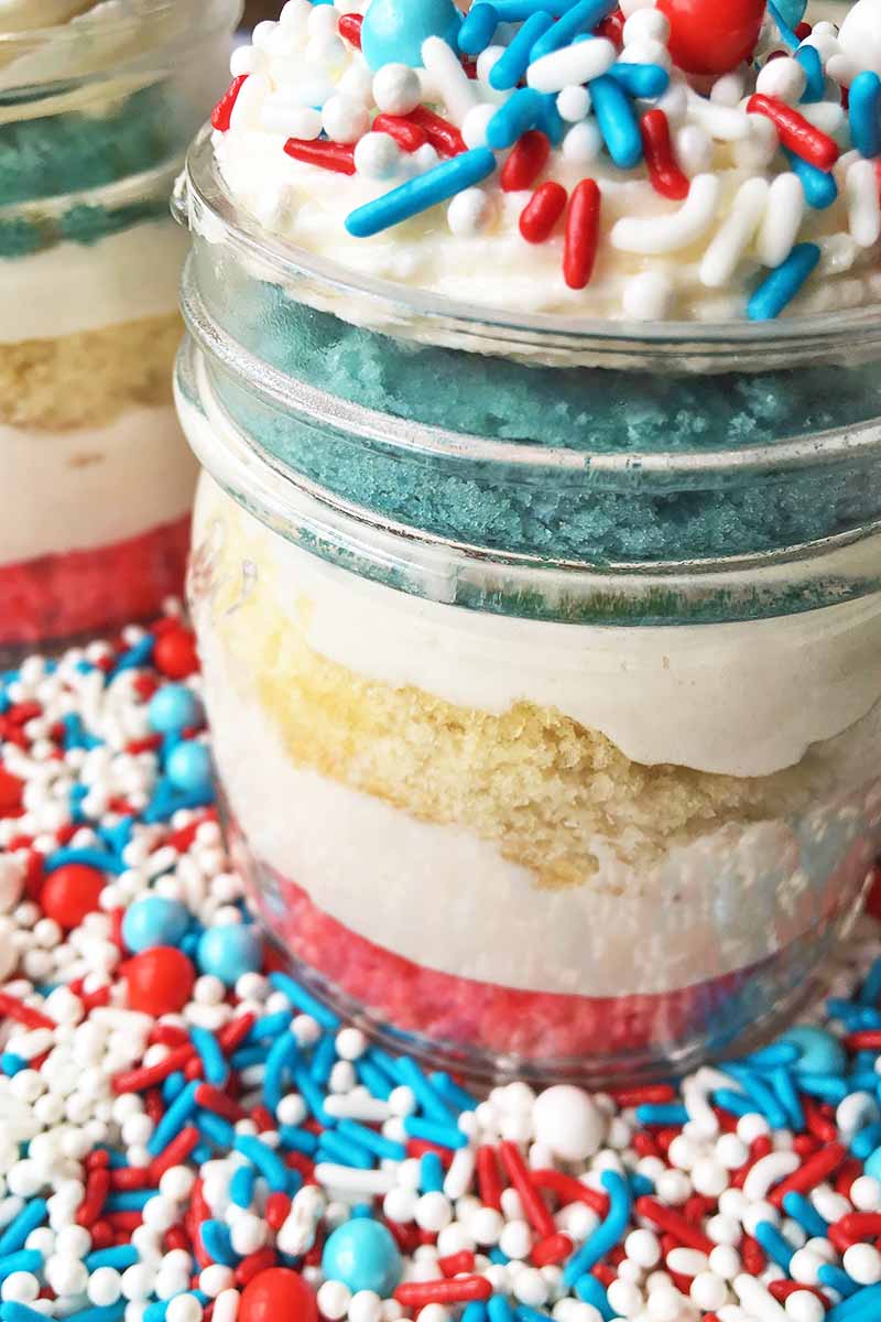 Vertical close-up image of a glass container filled with layers of colored pastries and white frosting with sprinkles.