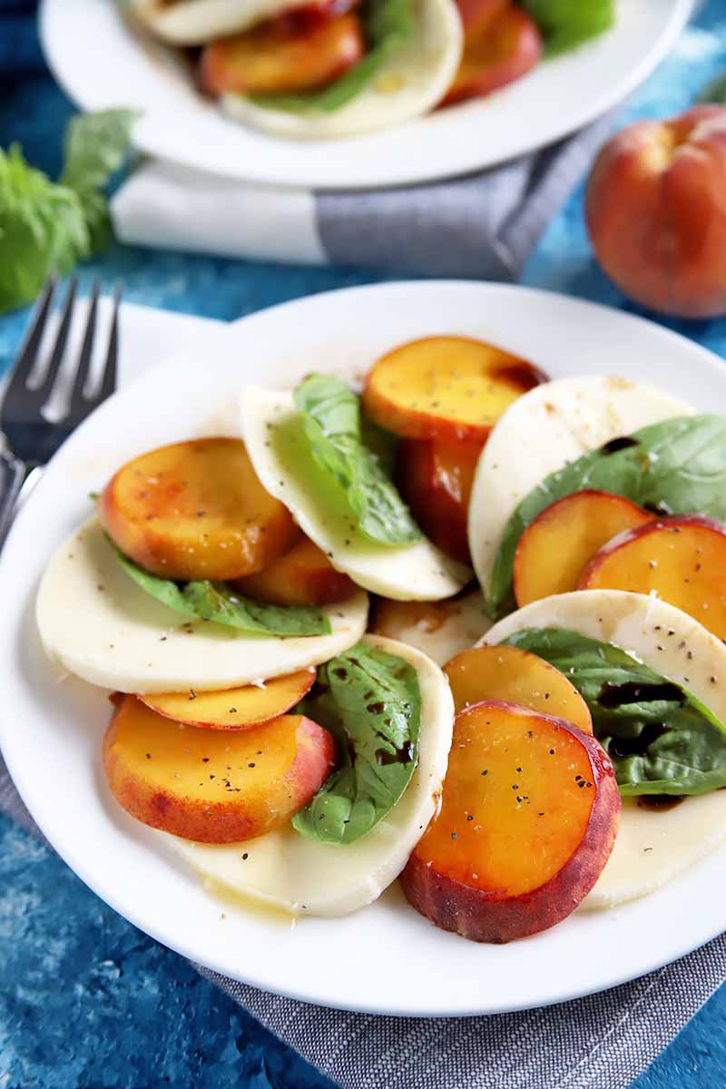 Vertical image of a white plate with sliced peaches, mozzarella, and basil covered in honey and vinegar next to a metal fork and napkin.