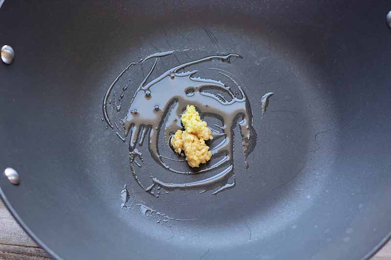 Horizontal image of a wok with oil, ginger, and garlic.