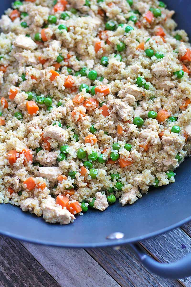 Vertical close-up image of a wok with fried quinoa, tofu, carrots, and peas.