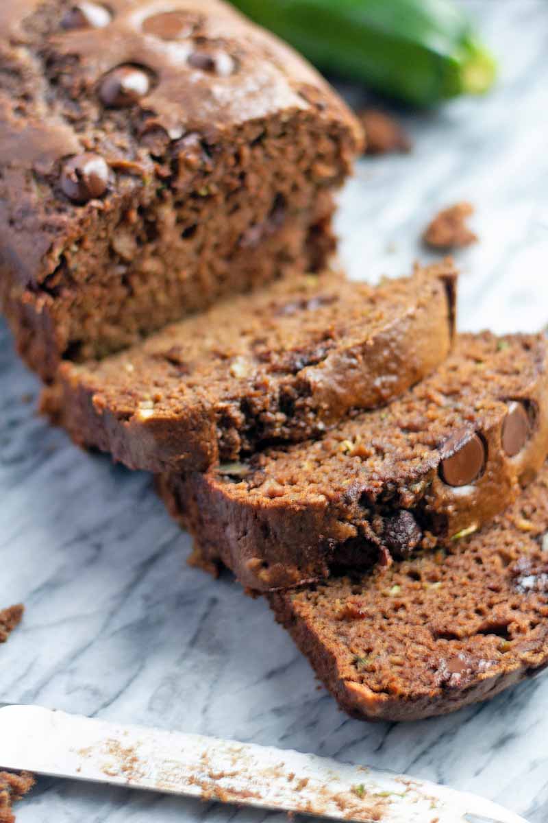 Vertical closeup image of a partially sliced loaf of chocolate quick bread on a marble surface, with a zucchini and a serrated knife.