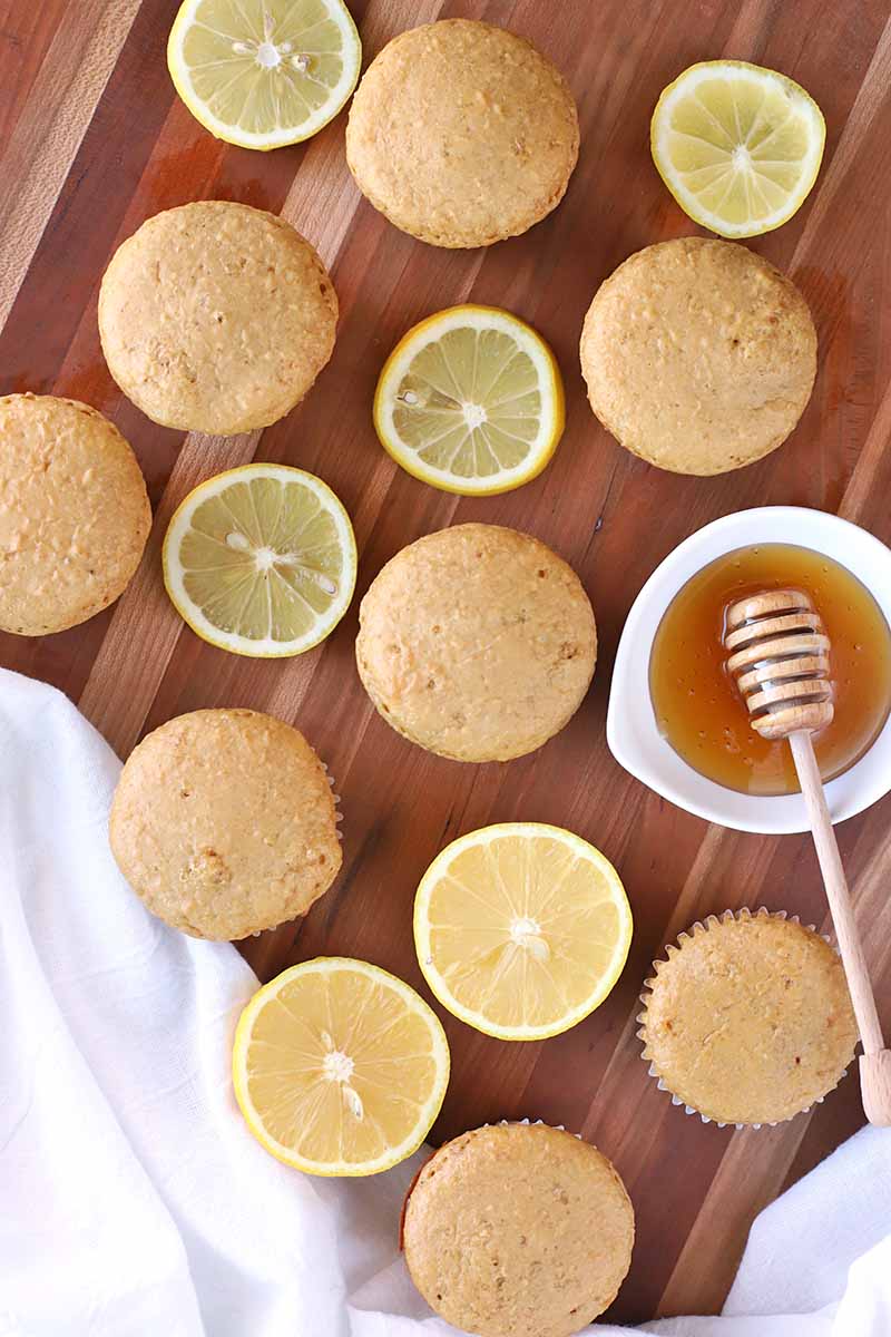 Vertical overhead image of muffins, lemon slices, a small ceramic dish of honey with a wooden dipper, and a white cloth on a brown wood surface with a few lighter beige stripes.