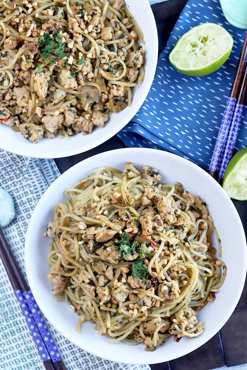 Vertical overhead image of two white ceramic bowls of gluten-free noodles and ground chicken, topped with cilantro and chopped peanuts, with sliced pieces of lime and two sets of chopsticks on two cloth napkins, on a dark brown wood surface.