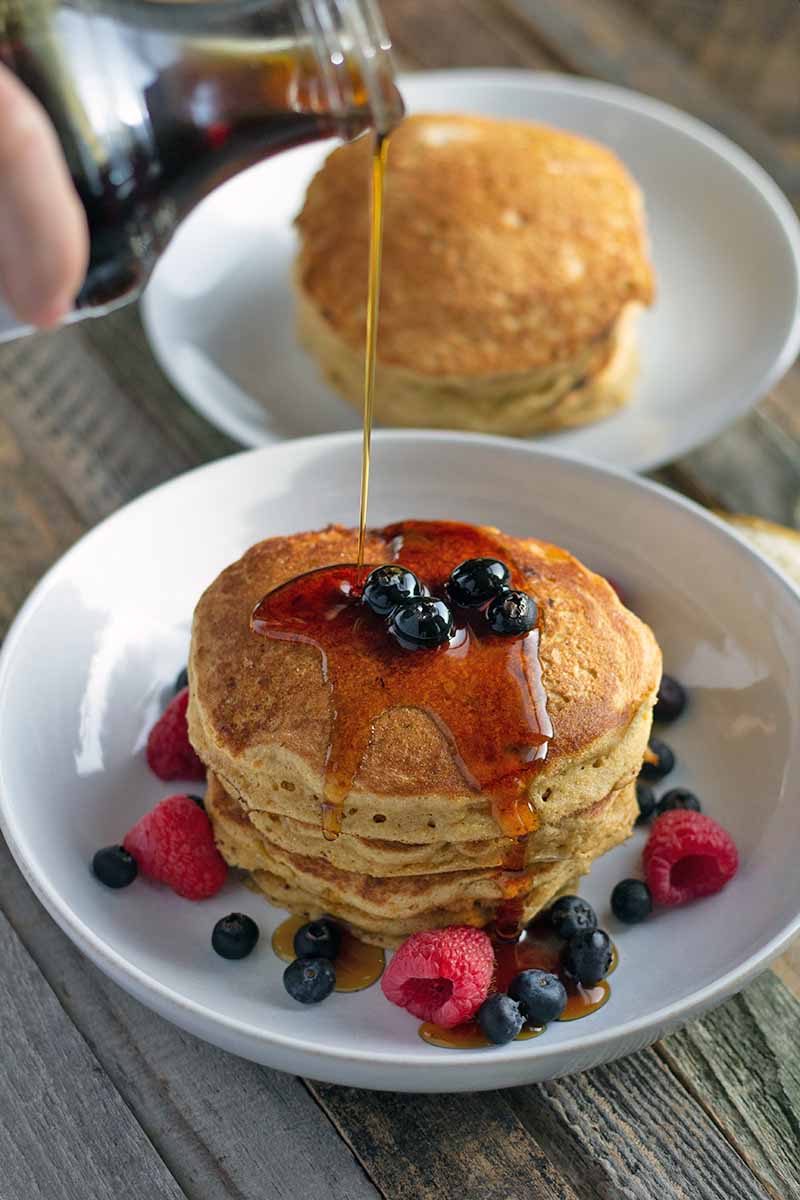 Vertical image of a maple syrup being poured from a glass bottle to the left of the frame onto a stack of pancakes in a white shallow bowl with fresh berries, with another plate of flapjacks in the background, on an unfinished wood table.
