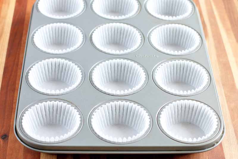 Horizontal image of a muffin tin with white paper liners arranged in each well, on a beige and brown striped wood surface.