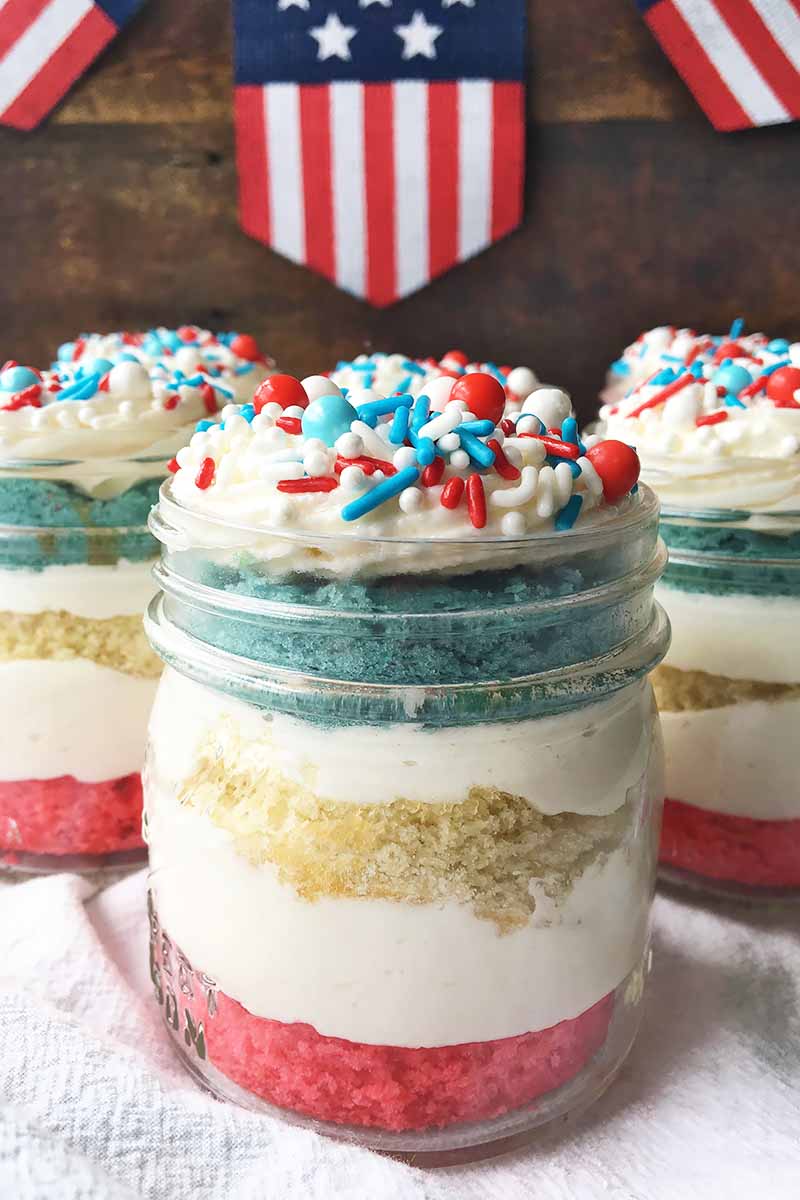 Vertical image of three trifles with layers of colored cakes, frosting, and sprinkles in front of flags.