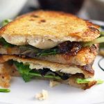 Horizontal image of two golden brown toasted halves of a sandwich with salad greens, fig jam, goat cheese, and chicken, on a white plate.