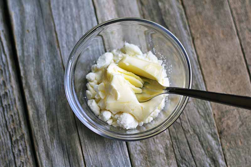 Overhead horizontal image of a small glass bowl of butter being mashed together with crumbled goat cheese with a fork, on a wood surface that is neither painted nor varnished.