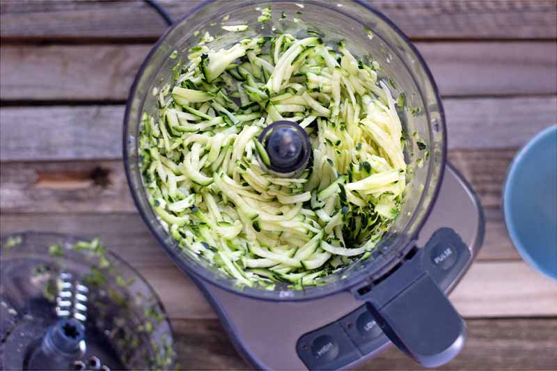 Horizontal overhead image of a food processor full of shredded zucchini with two bowls on an unfinished wood surface.