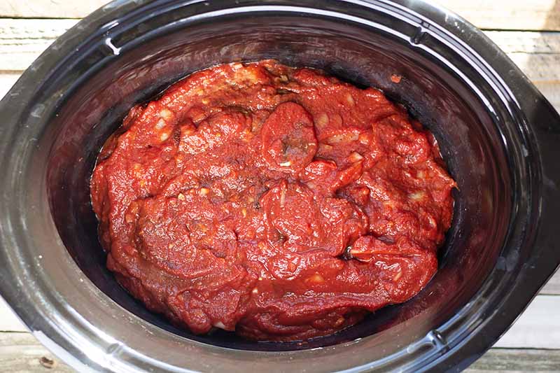 Overhead horizontal image of red sauce in the bottom of a slow cooker, on a wood surface.