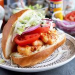 Horizontal image of a whole grilled shrimp po'boy with shredded lettuce, onions, and tomatoes with all of the ingredients in the background.