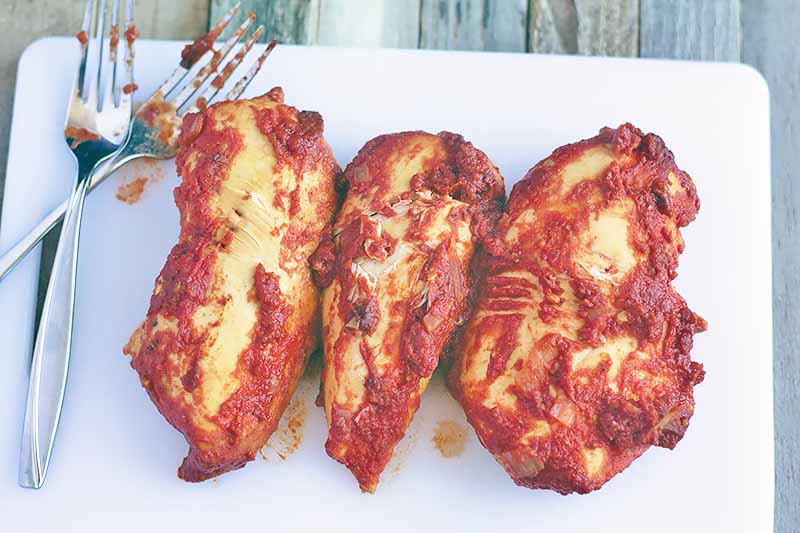 Horizontal image of three chicken breasts cooked in red sauce on a white cutting board with two forks, on a brown unfinished wood surface.