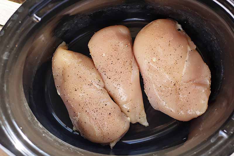 Horizontal overhead image of three uncooked boneless, skinless chicken breasts seasoned with salt and pepper in the bottom of a slow cooker.