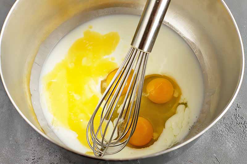 Horizontal image of a whisk in a pot with eggs, milk, and melted butter.