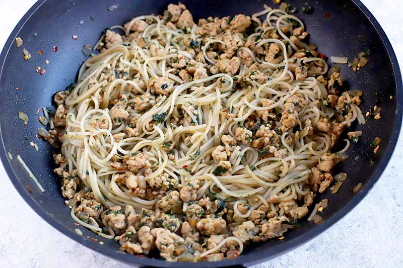 Overhead horizontal image of a wok filled with cooked noodles and ground chicken, a light brown sauce, and fresh herbs, on a white surface with light blue speckles.