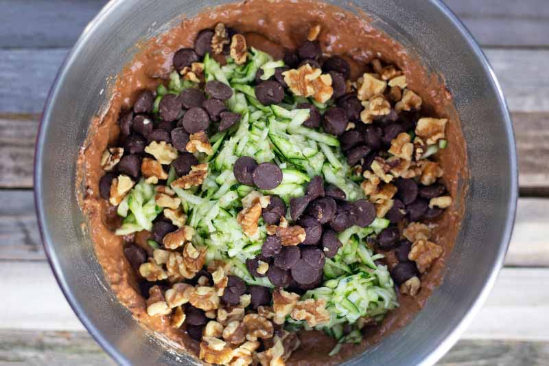 Horizontal overhead closely cropped image of a stainless steel mixing bowl containing batter with chocolate chips, shredded zucchini, and walnuts on top, on an unfinished wood surface.