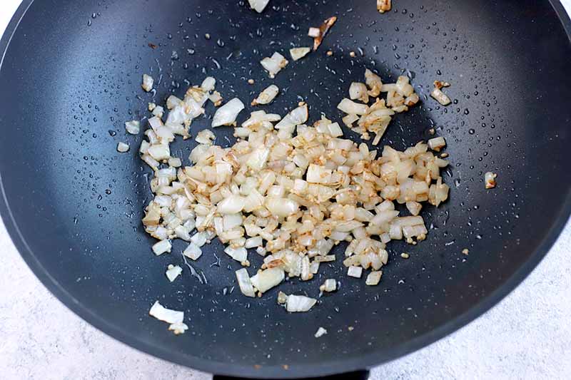 Overhead horizontal image of onions and garlic sauteeing in a black metal wok, on a white surface with light blue speckles.