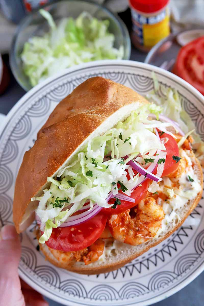 Vertical image of a hand holding a plate with a whole shrimp po'boy sandwich with fresh toppings.